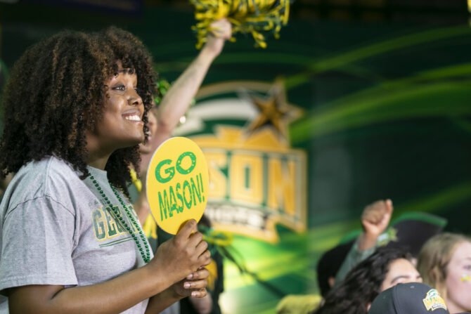 A woman holds a yellow “Go Mason!” sign while cheering at a George Mason University event, with green and gold decorations and other attendees in the background.