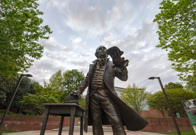 Statue of George Mason on the George Mason University campus, holding a scroll and standing beside a table with stacked books, with trees and campus buildings in the background.