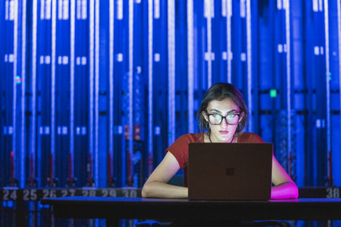 A woman sits at a table working on a laptop in front of a wall of blue lights in the background.