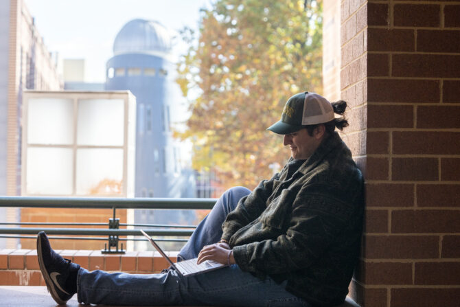 Student sitting on an outdoor balcony using a laptop, leaning against a brick wall. A campus building with a dome-shaped tower and autumn trees are visible in the background.