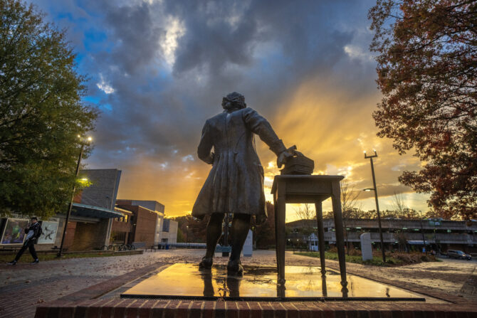 The George Mason statue on Fairfax campus at sunset, holding a scroll and positioned beside a table with stacked books. Sunlight streams through dramatic clouds behind the statue, with campus buildings and trees visible in the background.