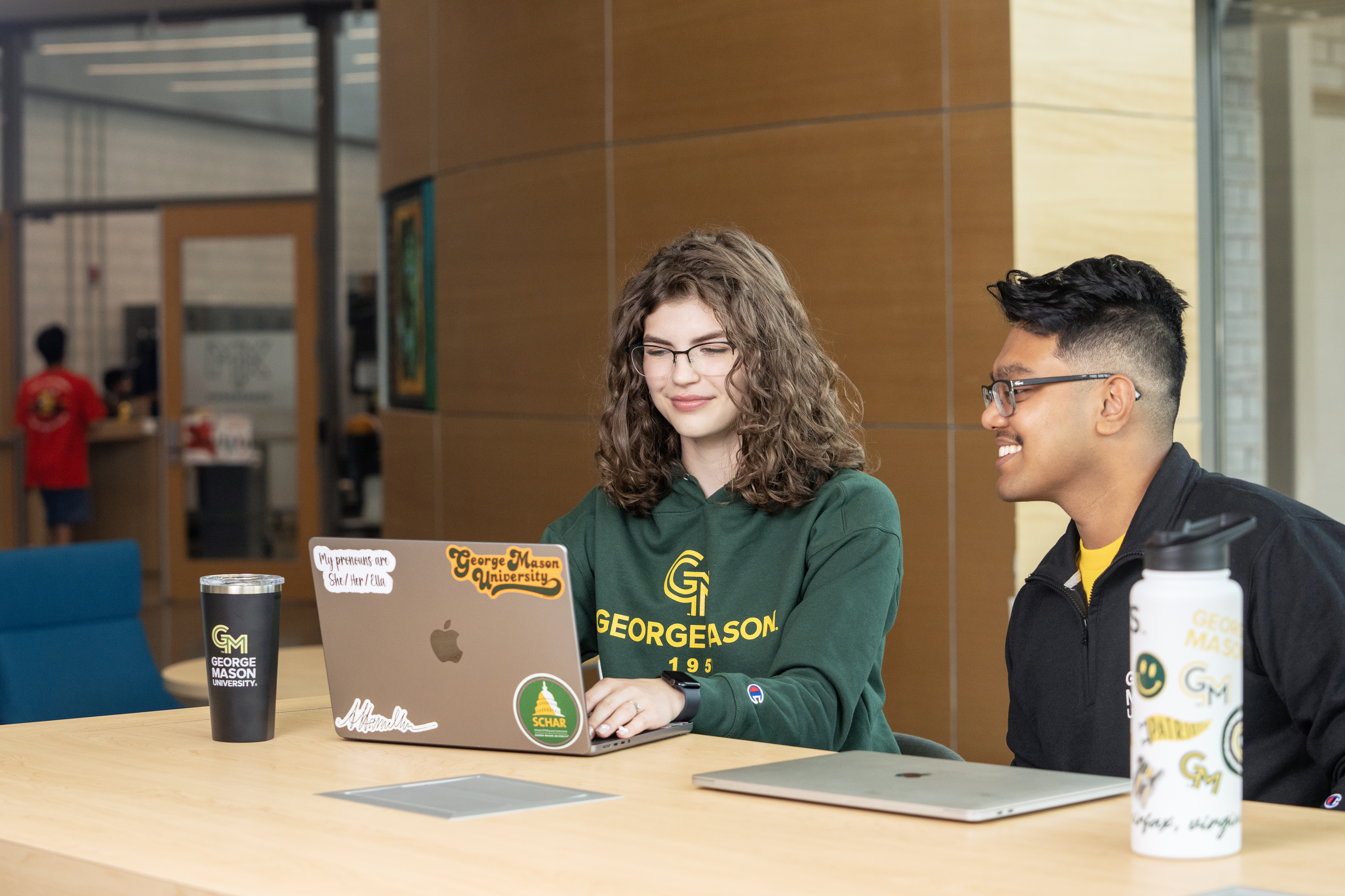 Two students sit together at a table in a campus building. They are both looking at a laptop on the table, which is covered with George Mason University stickers.