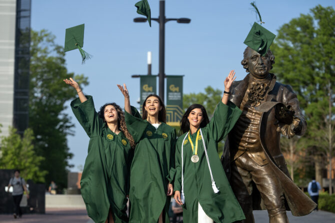 Three graduates in green caps and gowns toss their caps in the air beside a bronze George Mason statue on campus.