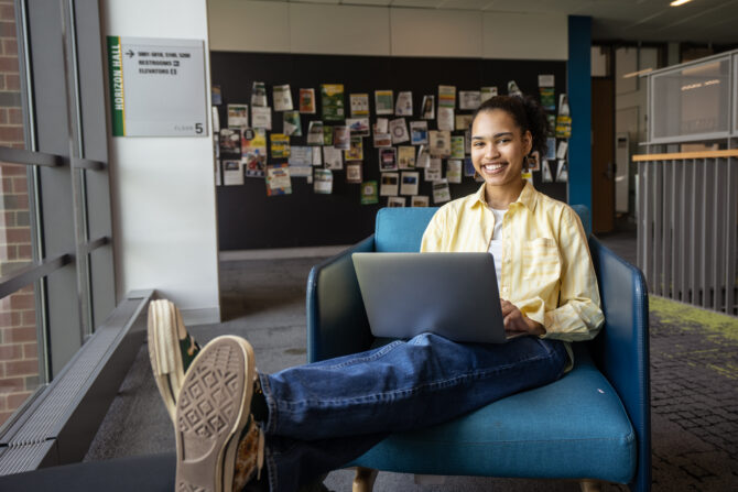 A student sits in a blue lounge chair using a laptop in a campus building. A bulletin board covered with flyers and brochures is visible in the background, and large windows line the study area.