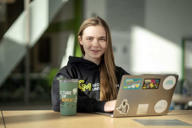 Student seated at a table using a laptop in a campus building.