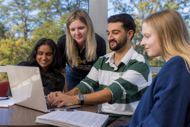 Four students collaborate in a campus study space, working together on a laptop near a window with trees visible outside